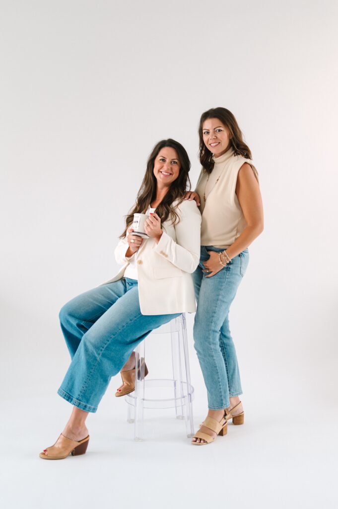 Two mortgage home loan professionals standing next to each other with a white background. One is standing and one is sitting on a stool holding a coffee mug.