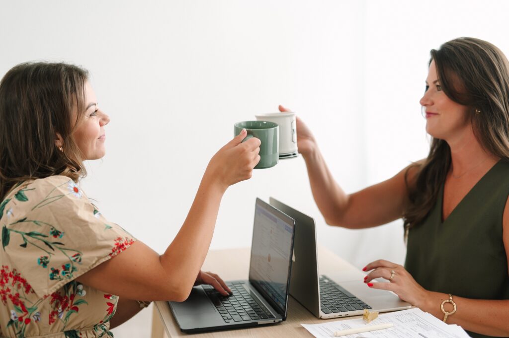 Detail photograph of two people clicking coffee mugs over their laptops.