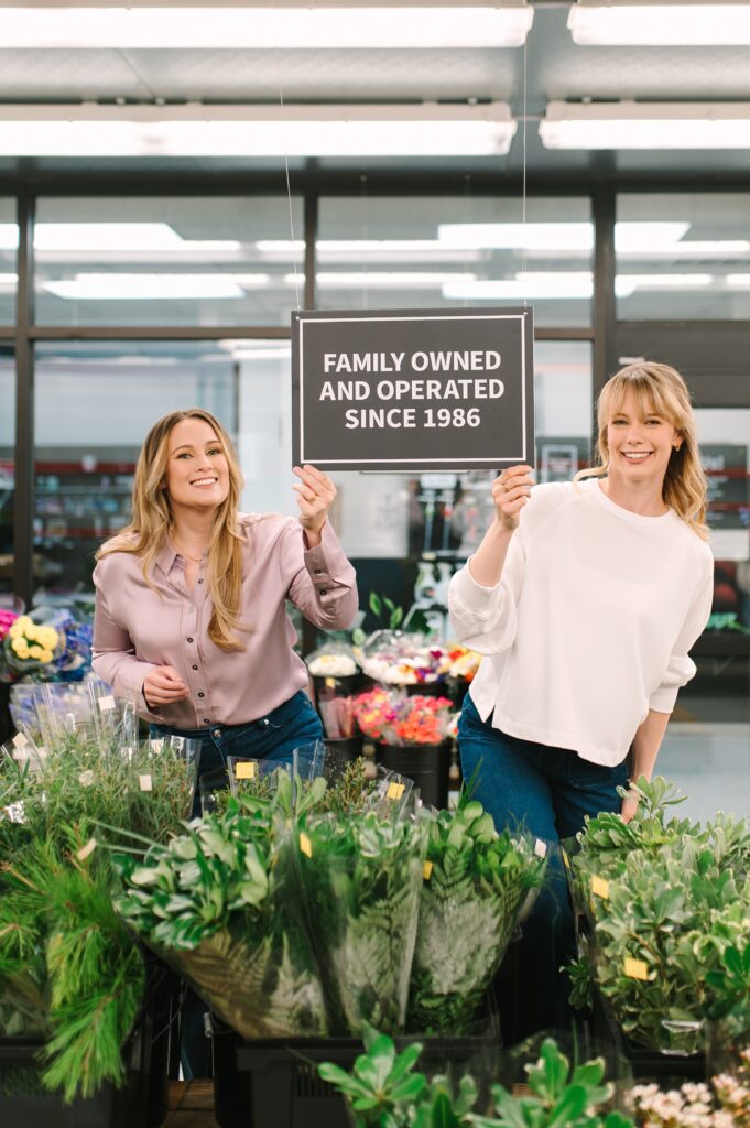 Two sisters at Wholesale Flower Market holding a Family Owned and Operated Since 1986 sign during branding photography.