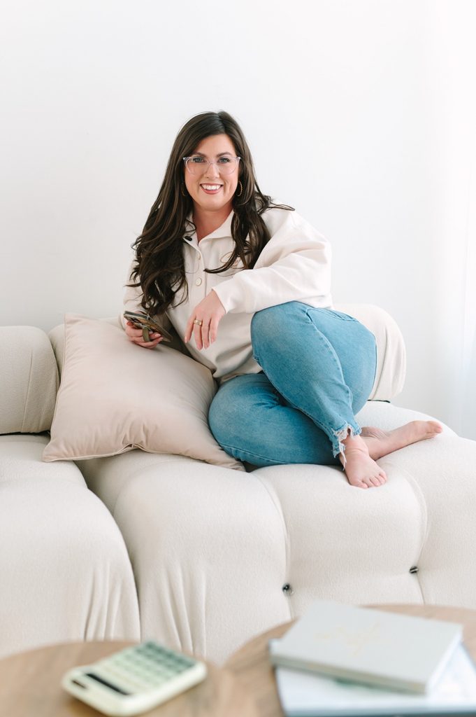 Smiling team member in a cream Sparrow Home Loans pullover and jeans sitting on a white sofa holding her phone