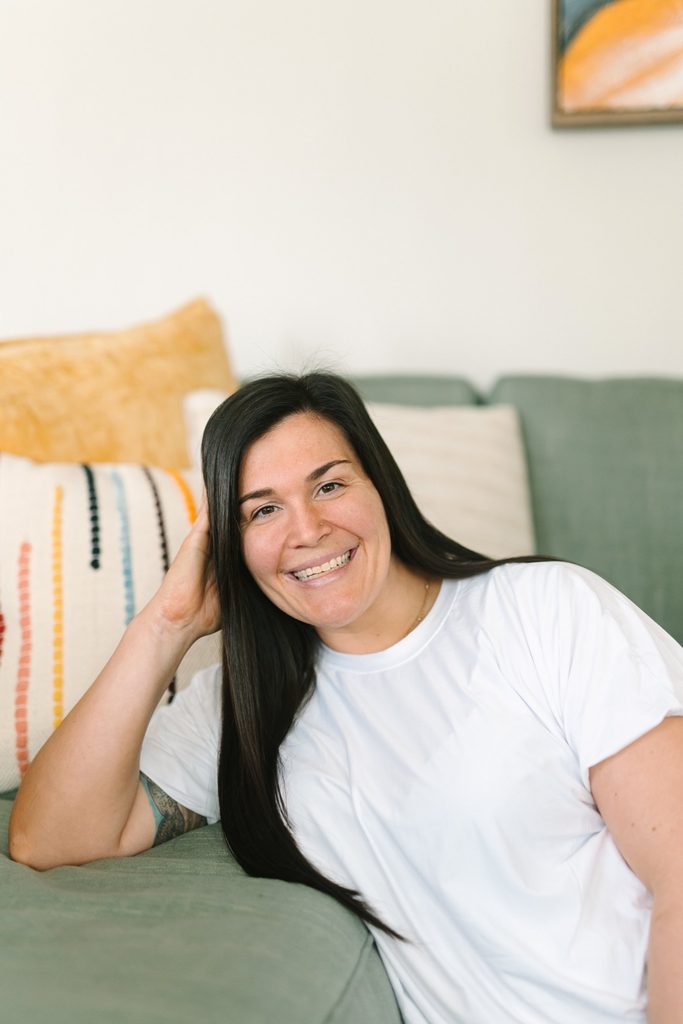 Smiling team member in a white tee leaning on a sage green sofa with colorful pillows, Richmond VA team photo