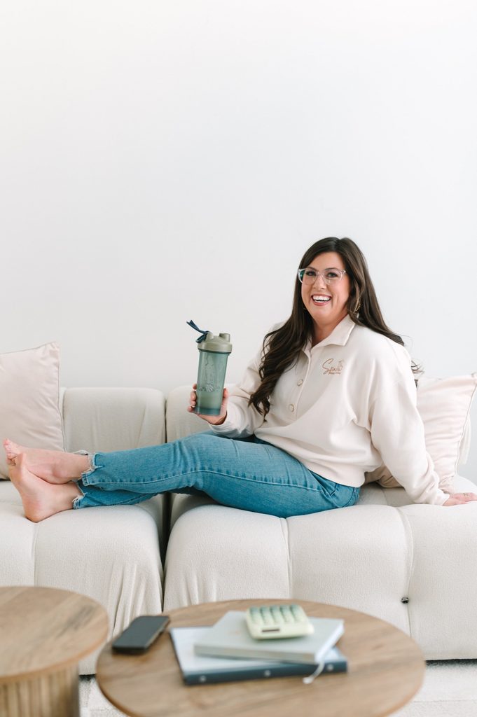 Laughing team member in a Sparrow Home Loans sweatshirt relaxing on a cream sofa with a shaker bottle, calculator and notebooks nearby
