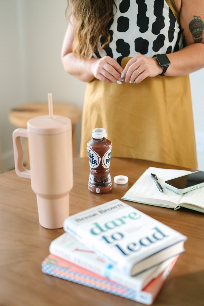 Desk details with a Dare to Lead book stack, pink tumbler, protein shake, open notebook and phone, Sparrow Home Loans branding