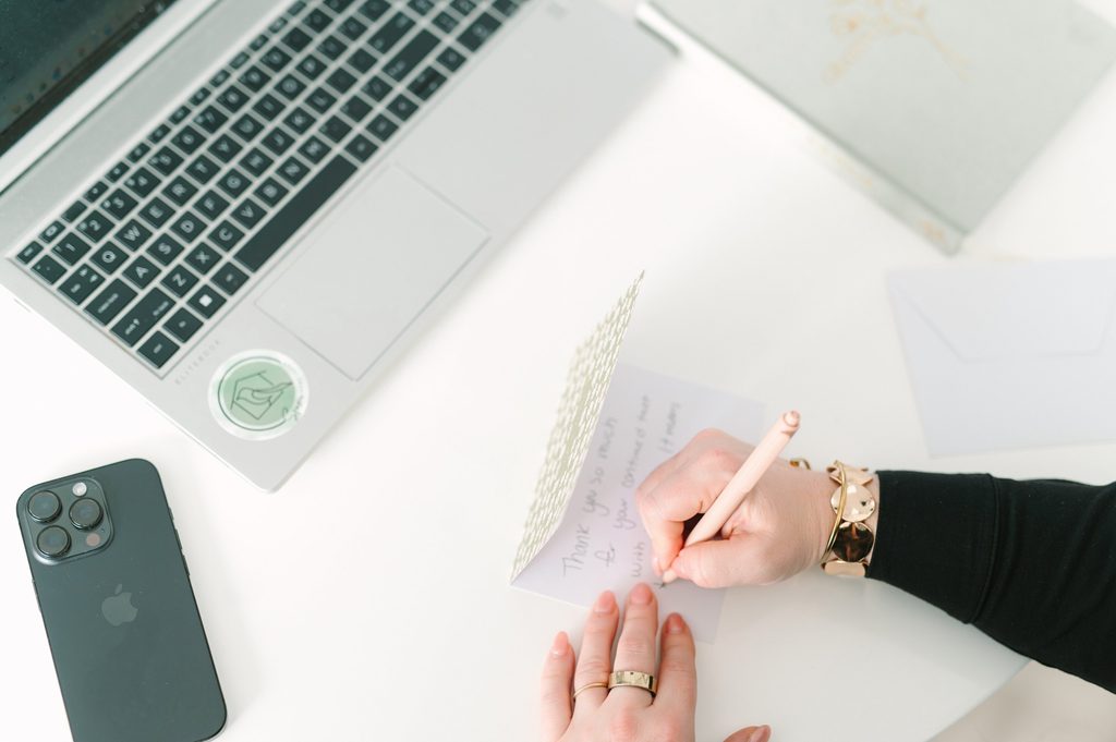Overhead view of a hand writing a thank-you card beside a laptop and phone on a white desk, Sparrow Home Loans social content